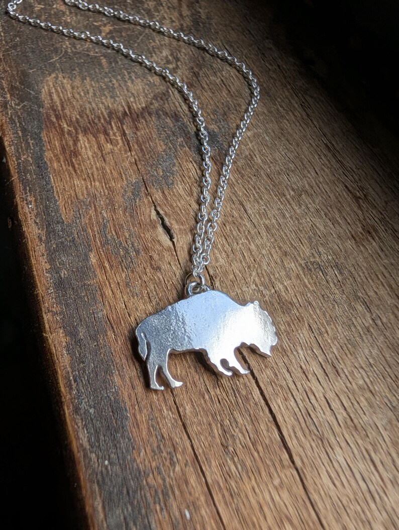 May include: A silver-toned metal pendant shaped like a buffalo silhouette, suspended from a matching silver-toned link chain. The pendant features a smooth, reflective surface and is attached to the chain by a small circular jump ring.