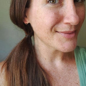 May include: A close-up of a woman's face with a small gold hoop earring in her left ear. She has long brown hair pulled back in a ponytail and is wearing a green shirt.