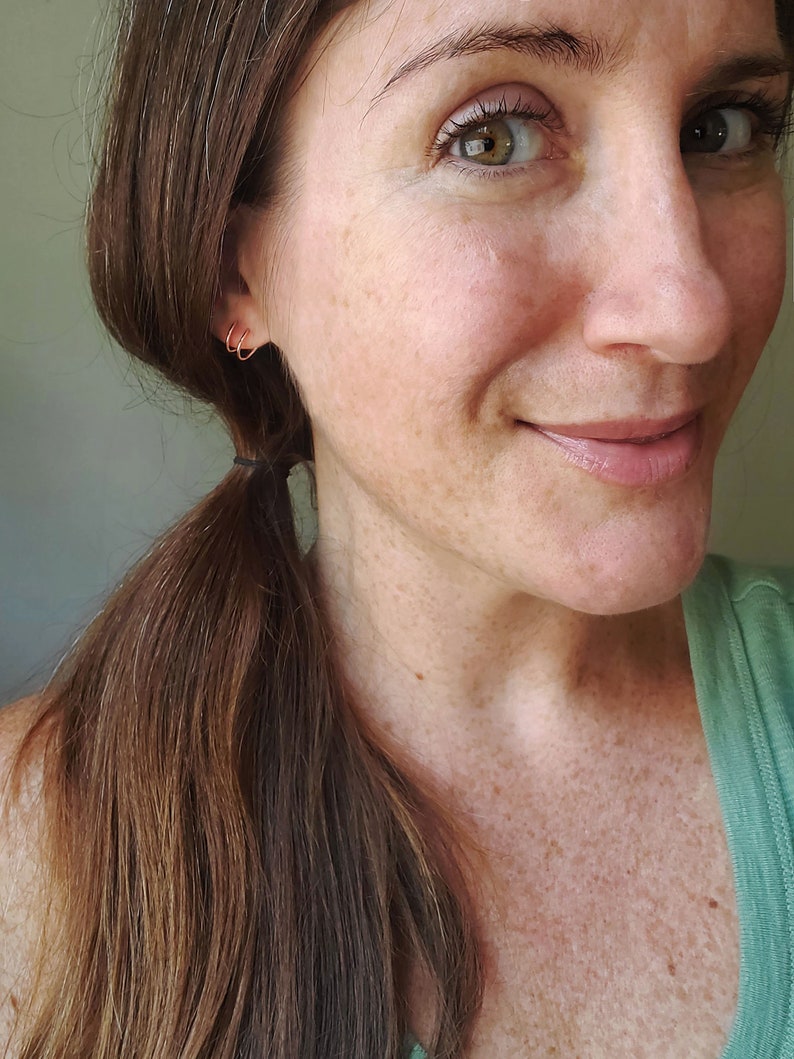 May include: A close-up of a woman's face with a small gold hoop earring in her left ear. She has long brown hair pulled back in a ponytail and is wearing a green shirt.