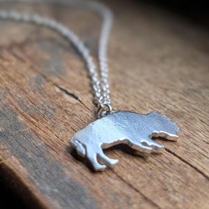 May include: A silver-toned metal pendant shaped like a buffalo silhouette, suspended from a matching delicate silver-toned link chain. The pendant features a smooth, reflective surface and is attached to the chain by a small circular jump ring.