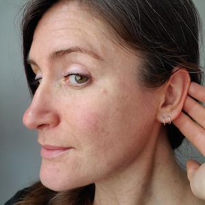 May include: A woman with brown hair and a light complexion is shown in profile. She is wearing a black shirt and has her hand to her ear. She is wearing two small silver hoop earrings in her left ear.