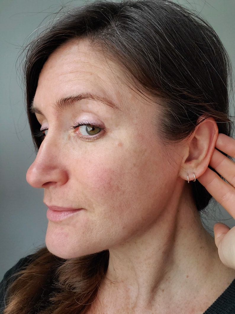 May include: A woman with brown hair and a light complexion is shown in profile. She is wearing a black shirt and has her hand to her ear. She is wearing two small silver hoop earrings in her left ear.