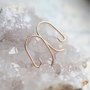 May include: A pair of gold-toned metal earrings featuring a smooth, continuous wire bent into an elongated, open-ended oval shape. The earrings have a minimalist, curved design with rounded ends, showcasing a polished metallic finish throughout.