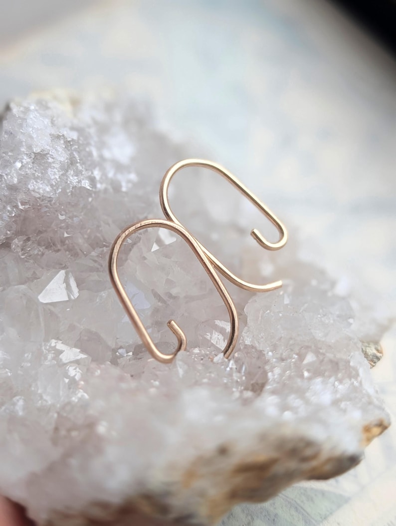 May include: A pair of gold-toned metal earrings featuring a smooth, continuous wire bent into an elongated, open-ended oval shape. The earrings have a minimalist, curved design with rounded ends, showcasing a polished metallic finish throughout.