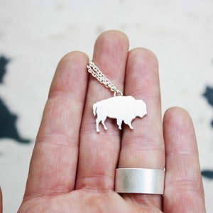 May include: A silver-colored necklace featuring a flat, silhouette-style pendant shaped like a bison. The pendant is attached to a delicate silver-colored chain. A wide, plain silver-colored band ring is also visible on the finger.