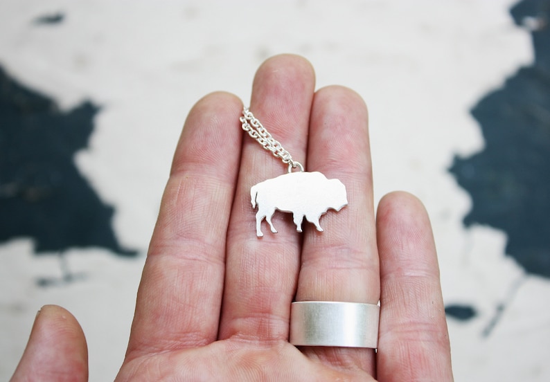 May include: A silver-colored necklace featuring a flat, silhouette-style pendant shaped like a bison. The pendant is attached to a delicate silver-colored chain. A wide, plain silver-colored band ring is also visible on the finger.