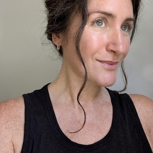 May include: A small, thin, gold-toned rectangular hoop earring worn in the earlobe. The earring features a minimalist, geometric shape with a polished metallic finish, designed as a simple, lightweight loop that secures through the ear piercing.