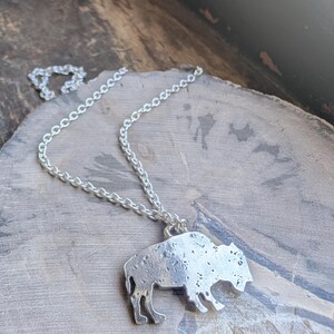 May include: A silver-toned metal pendant shaped like a bison, featuring a textured, mottled surface finish. The pendant is attached to a simple silver-toned link chain that loops around the top of the bison figure, creating a necklace.