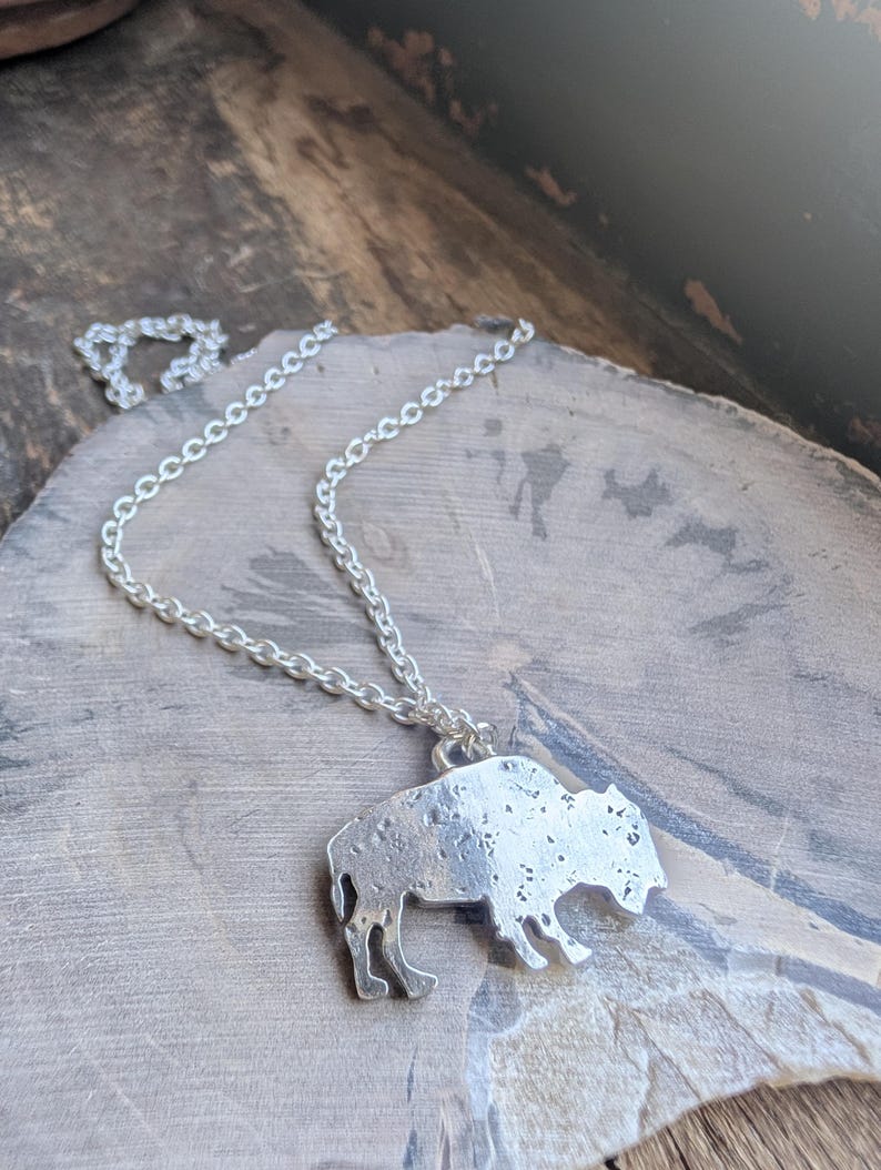 May include: A silver-toned metal pendant shaped like a bison, featuring a textured, mottled surface finish. The pendant is attached to a simple silver-toned link chain that loops around the top of the bison figure, creating a necklace.