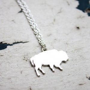 May include: A silver-toned necklace featuring a flat, solid pendant shaped like a bison in profile. The pendant is attached to a delicate, fine-link silver-toned chain that extends upwards from the top of the bison's back, creating a simple piece.