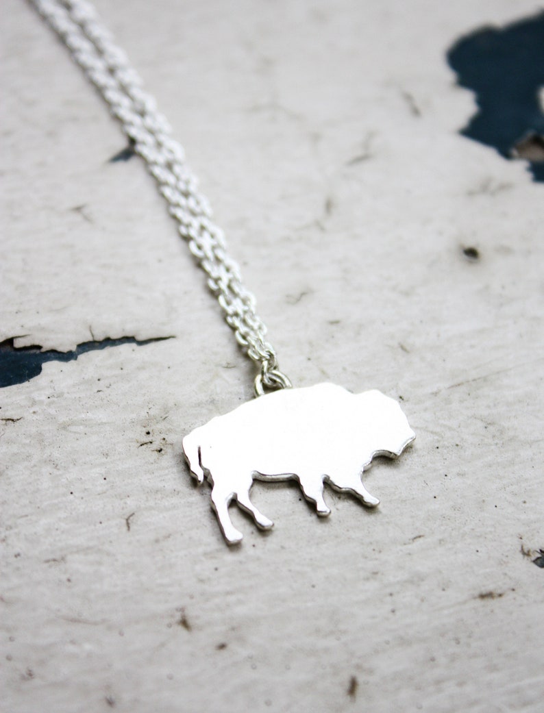 May include: A silver-toned necklace featuring a flat, solid pendant shaped like a bison in profile. The pendant is attached to a delicate, fine-link silver-toned chain that extends upwards from the top of the bison's back, creating a simple piece.