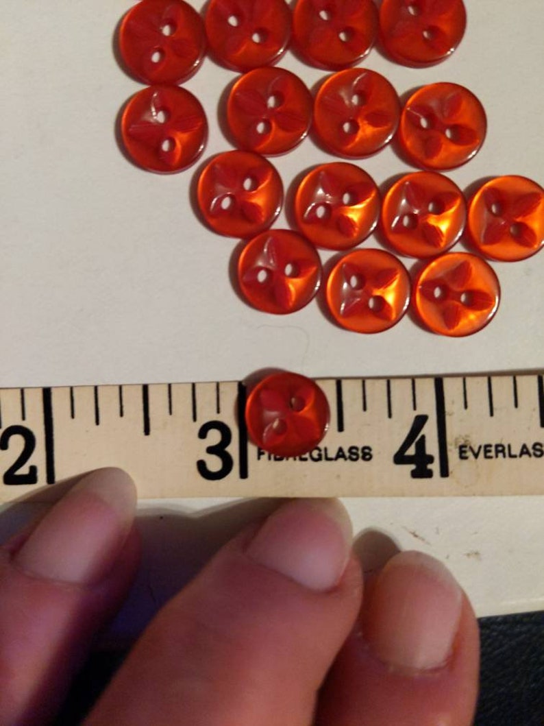 Puede incluir: Colecci&oacute;n de botones rojos transl&uacute;cidos con dise&ntilde;o de cuatro p&eacute;talos. Cada bot&oacute;n tiene dos agujeros y mide aproximadamente 2,5 cm de di&aacute;metro. Los botones est&aacute;n dispuestos sobre una superficie blanca.