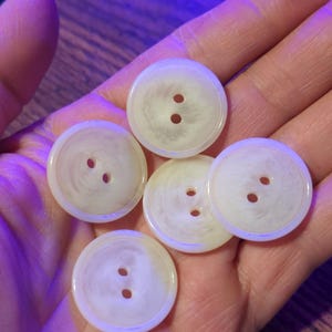 May include: A collection of six off-white, round buttons with two holes each. The buttons have a subtle, marbled appearance and a slightly glossy finish. These buttons are ideal for sewing projects and garment construction.