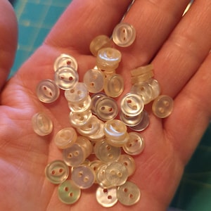 May include: A collection of white pearl-like buttons with two holes each. The buttons are arranged in a pile on a hand.