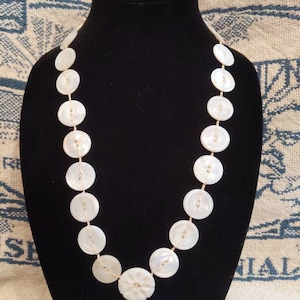 May include: A white button necklace with gold thread. The buttons are round and have a slightly raised center. The necklace is on a black mannequin.