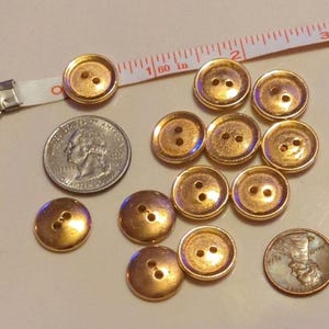 Puede incluir: Una colecci&oacute;n de 12 botones de color dorado, cada uno con dos agujeros. Los botones est&aacute;n dispuestos en semic&iacute;rculo sobre una superficie blanca. Se incluye una moneda de 25 centavos y un centavo como referencia de tama&ntilde;o.