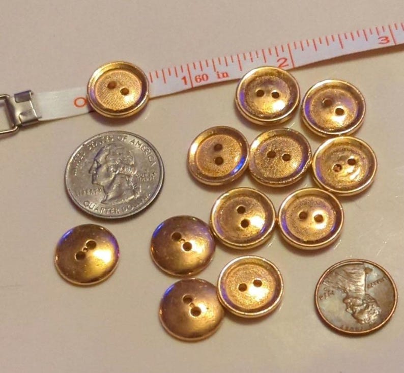 Puede incluir: Una colecci&oacute;n de 12 botones de color dorado, cada uno con dos agujeros. Los botones est&aacute;n dispuestos en semic&iacute;rculo sobre una superficie blanca. Se incluye una moneda de 25 centavos y un centavo como referencia de tama&ntilde;o.
