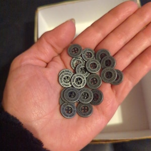 May include: A collection of small, silver-colored buttons with the Harley-Davidson logo. The buttons are round with a raised edge and a central hole. The text "HARLEY-DAVIDSON" is inscribed around the edge of each button. The buttons are held in a person's hand.