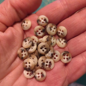 May include: A collection of brown and white speckled buttons with four holes each. The buttons are arranged in a pile in a person's hand.