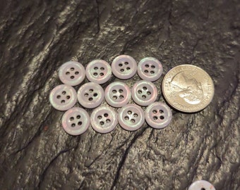 May include: A collection of 14 white buttons with a iridescent finish and four holes. The buttons have a subtle pink and green color shift. The buttons are arranged in a circle around a US quarter.