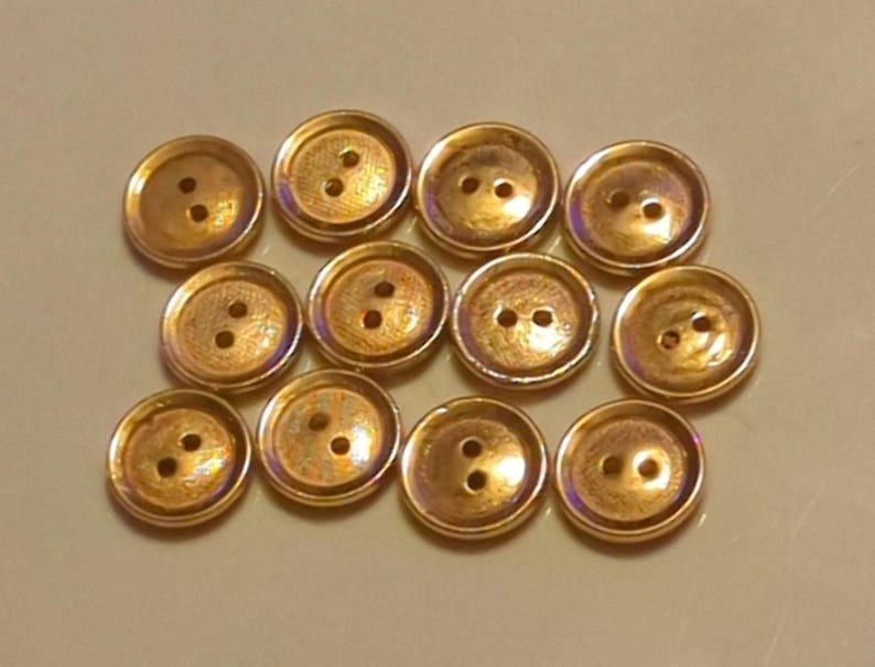 Puede incluir: Una colecci&oacute;n de 10 botones de color dorado con dos agujeros cada uno. Los botones tienen un acabado ligeramente iridiscente.