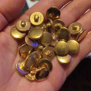 May include: A pile of gold-colored metal buttons with a shank on the back. The buttons are round and have a smooth, shiny finish.