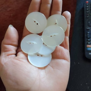 May include: A collection of six round, off-white buttons with two small holes for sewing. The buttons are held in a hand, showcasing their smooth, slightly translucent appearance. These buttons are ideal for sewing projects.