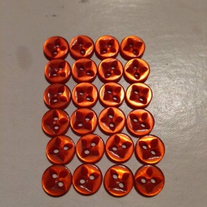 Puede incluir: Veinticuatro botones naranjas dispuestos en una cuadr&iacute;cula. Cada bot&oacute;n tiene un dise&ntilde;o de cuatro p&eacute;talos y cuatro agujeros para coser. Los botones son redondos y tienen un acabado brillante, ideales para proyectos de costura.