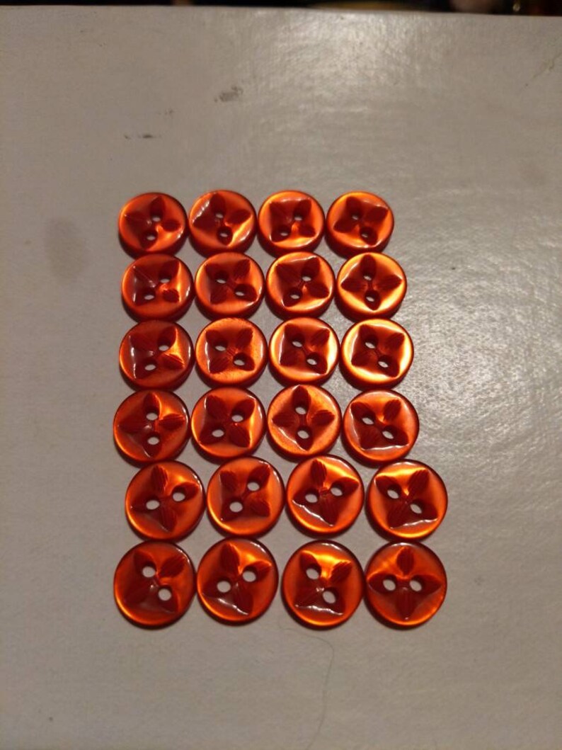 Puede incluir: Veinticuatro botones naranjas dispuestos en una cuadr&iacute;cula. Cada bot&oacute;n tiene un dise&ntilde;o de cuatro p&eacute;talos y cuatro agujeros para coser. Los botones son redondos y tienen un acabado brillante, ideales para proyectos de costura.