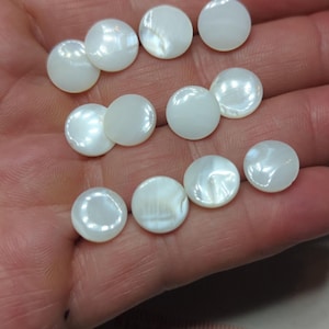 May include: A collection of ten round, white mother-of-pearl buttons with a smooth, iridescent finish. The buttons are arranged in a cluster, and a portion of a measuring tape showing numbers 7, 8, and 9 in centimetres is visible at the bottom.