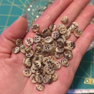 May include: A handful of small, round buttons with four holes each. The buttons have a mottled pattern of cream and brown, with a slightly glossy finish. These buttons are ideal for sewing and craft projects.