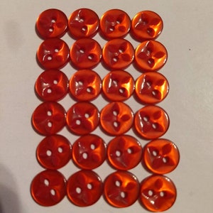 Puede incluir: Veinticuatro botones naranjas dispuestos en una cuadr&iacute;cula. Cada bot&oacute;n tiene un dise&ntilde;o de cuatro p&eacute;talos y dos agujeros para coser. Los botones tienen un acabado brillante y son de un tono naranja vibrante.