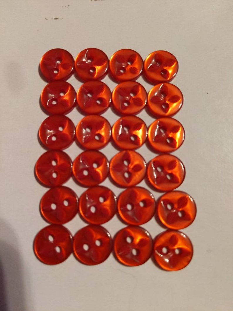 Puede incluir: Veinticuatro botones naranjas dispuestos en una cuadr&iacute;cula. Cada bot&oacute;n tiene un dise&ntilde;o de cuatro p&eacute;talos y dos agujeros para coser. Los botones tienen un acabado brillante y son de un tono naranja vibrante.