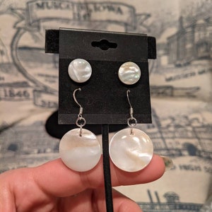 May include: A pair of white mother of pearl earrings with silver hooks. The earrings are round and have a subtle iridescent sheen. The earrings are displayed on a black card.