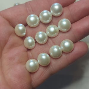 May include: A selection of creamy white pearls, each with a unique shape and size. The pearls are smooth and reflect light, creating a subtle shimmer. The pearls are held in a hand, highlighting their potential for crafting jewellery or other decorative items.