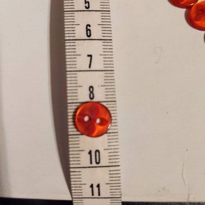 Puede incluir: Primer plano de un bot&oacute;n rojo transparente con dos agujeros, de aproximadamente 2 cm de di&aacute;metro, junto a una cinta m&eacute;trica. Varios botones m&aacute;s son visibles en el fondo. Los botones son probablemente para coser o manualidades.