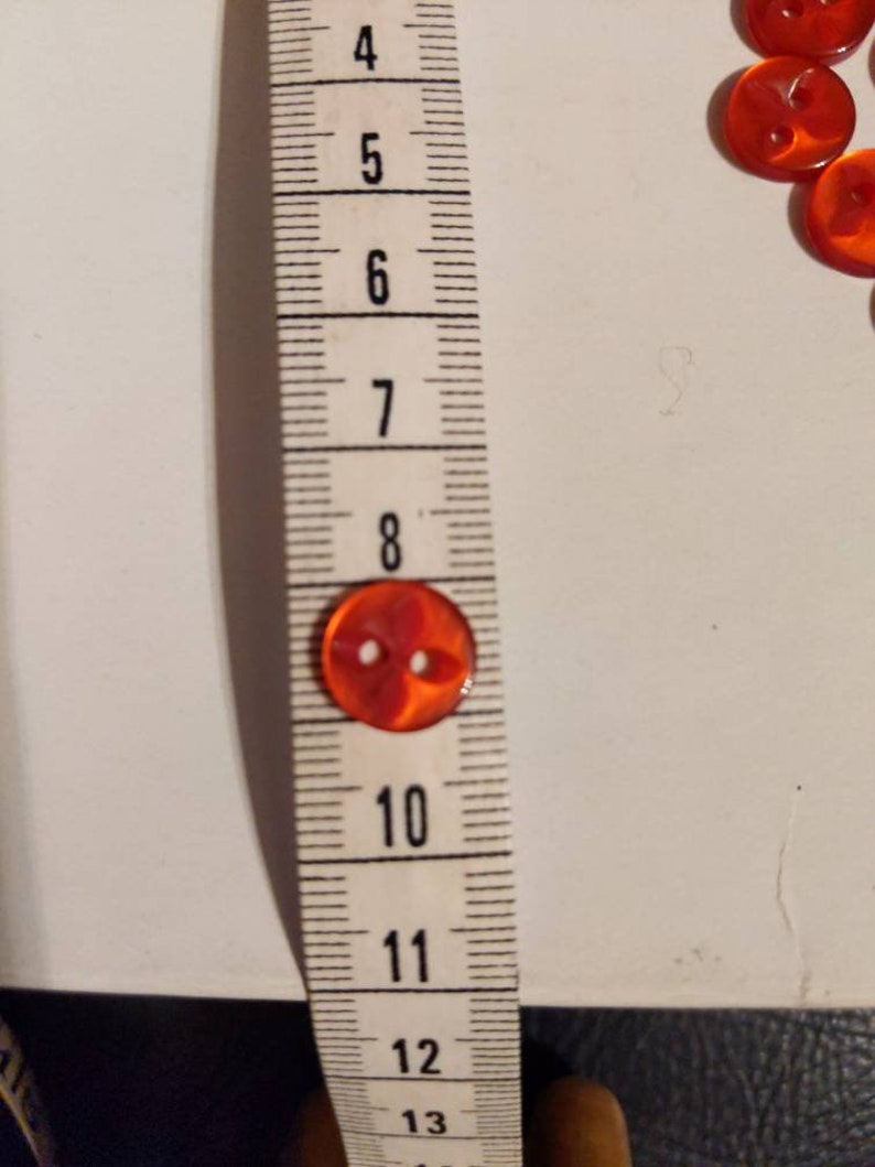 Puede incluir: Primer plano de un bot&oacute;n rojo transparente con dos agujeros, de aproximadamente 2 cm de di&aacute;metro, junto a una cinta m&eacute;trica. Varios botones m&aacute;s son visibles en el fondo. Los botones son probablemente para coser o manualidades.