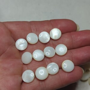 May include: A collection of eleven round, white buttons with a smooth, iridescent finish. Each button features a slightly domed surface that reflects light, showcasing the natural pearlescent sheen characteristic of shell material in a circular shape.