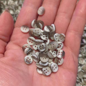 May include: A pile of small, white and gray buttons with four holes each. The buttons are made of a smooth, shiny material and have a mottled pattern.