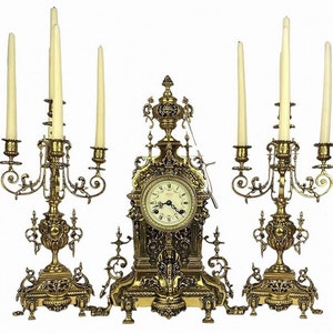 May include: A gold-toned clock and candle holder set. The clock has a white face with black Roman numeral hour markers and black hands. The candle holders have three candle arms each, with white candles.