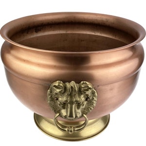 Könnte beinhalten: Ein Kupfertopf mit einem Löwenkopfdesign an den Griffen. Der Topf hat einen runden Boden und eine breite, flache Schale.