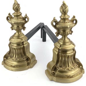 Peut inclure: Deux chenets de cheminée anciens en laiton avec un motif d'urne décorative et un support en métal noir. Les chenets présentent un design orné et détaillé avec un sommet en forme de flamme. Un élément de décoration intérieure.