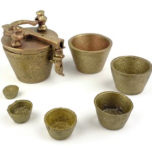 Puede incluir: Un conjunto de seis recipientes de metal de color dorado con un recipiente con tapa en el centro. Los recipientes son de diferentes tamaños y formas, con una superficie texturizada.