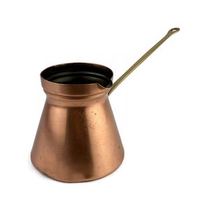 Puede incluir: Una cafetera de cobre con un mango de latón. La cafetera tiene un fondo redondeado y una parte superior acampanada.