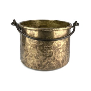 Op de afbeelding: Een gehamerde koperen pot met een zwart metalen handvat. De pot heeft een rustieke, antieke uitstraling.