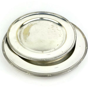 May include: Two silver serving plates, one stacked on top of the other. The top plate has a decorative border and the bottom plate has a more intricate design.