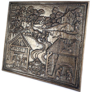 Peut inclure: Une plaque murale en métal avec un relief en relief représentant une scène rurale avec une église, des maisons et des personnages. La plaque est peinte en bronze et possède une bordure décorative. Le texte "Ora et Labora" est inscrit sur la plaque.