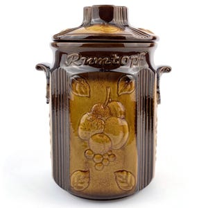 Könnte beinhalten: Brauner Keramik-Rumtopf mit Deckel. Die Dose zeigt geprägte Obst- und Blattmotive, mit dem Wort "Rumtopf" auf der Vorderseite. Sie hat zwei seitliche Griffe und eine dekorative Oberseite.