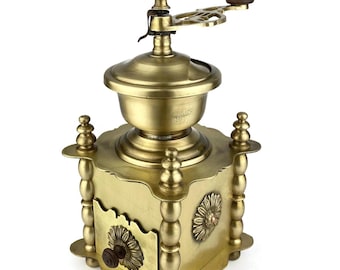 May include: A vintage brass coffee grinder with a wooden handle. The grinder features a square base with decorative floral accents and a crank mechanism on top. The brass has a polished finish, and the design is ornate.