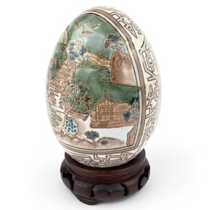 Może przedstawiać: Ozdobne jajo z ręcznie malowaną sceną krajobrazu w odcieniach zieleni, błękitu i brązu. Jajo jest umieszczone na ciemnej drewnianej podstawce. Projekt obejmuje pagody, drzewa i ptaki, z wzorzystą obwódką.